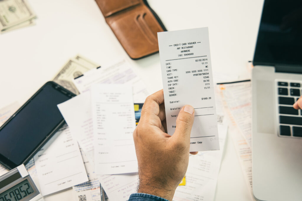 Mão segura comprovante de pagamento com cartão de crédito sobre mesa com notas fiscais, cédulas, carteira, celular, calculadora e notebook.
